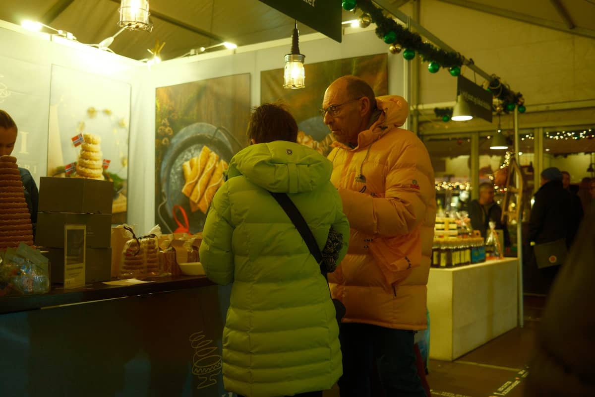 People in winter jackets order food at a brightly-lit indoor Christmas market stall selling pastries and sweets.