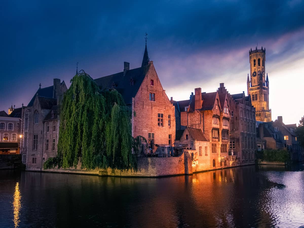 Evening view of Bruges' iconic Rozenhoedkaai canal and Belfry tower illuminated, with medieval buildings reflecting in the water under a dramatic sky.