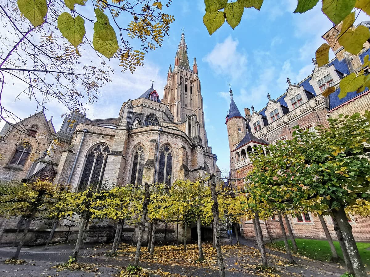 View of the Church of Our Lady in Bruges, featuring the tall brick Gothic tower and ornate apse framed by autumn trees and adjacent historic buildings.