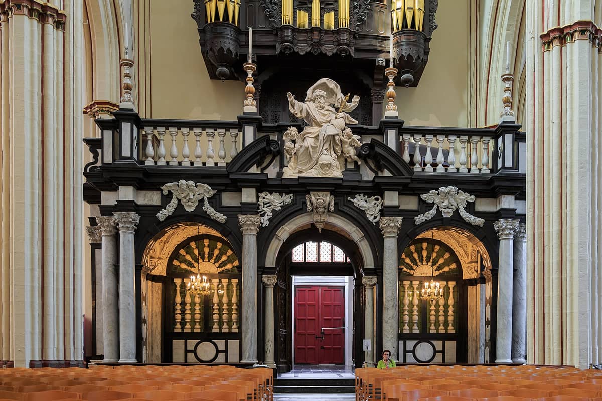 Interior view of Sint-Salvatorskathedraal in Bruges showing ornate organ loft, marble columns, sculpted figures, and wooden pews.