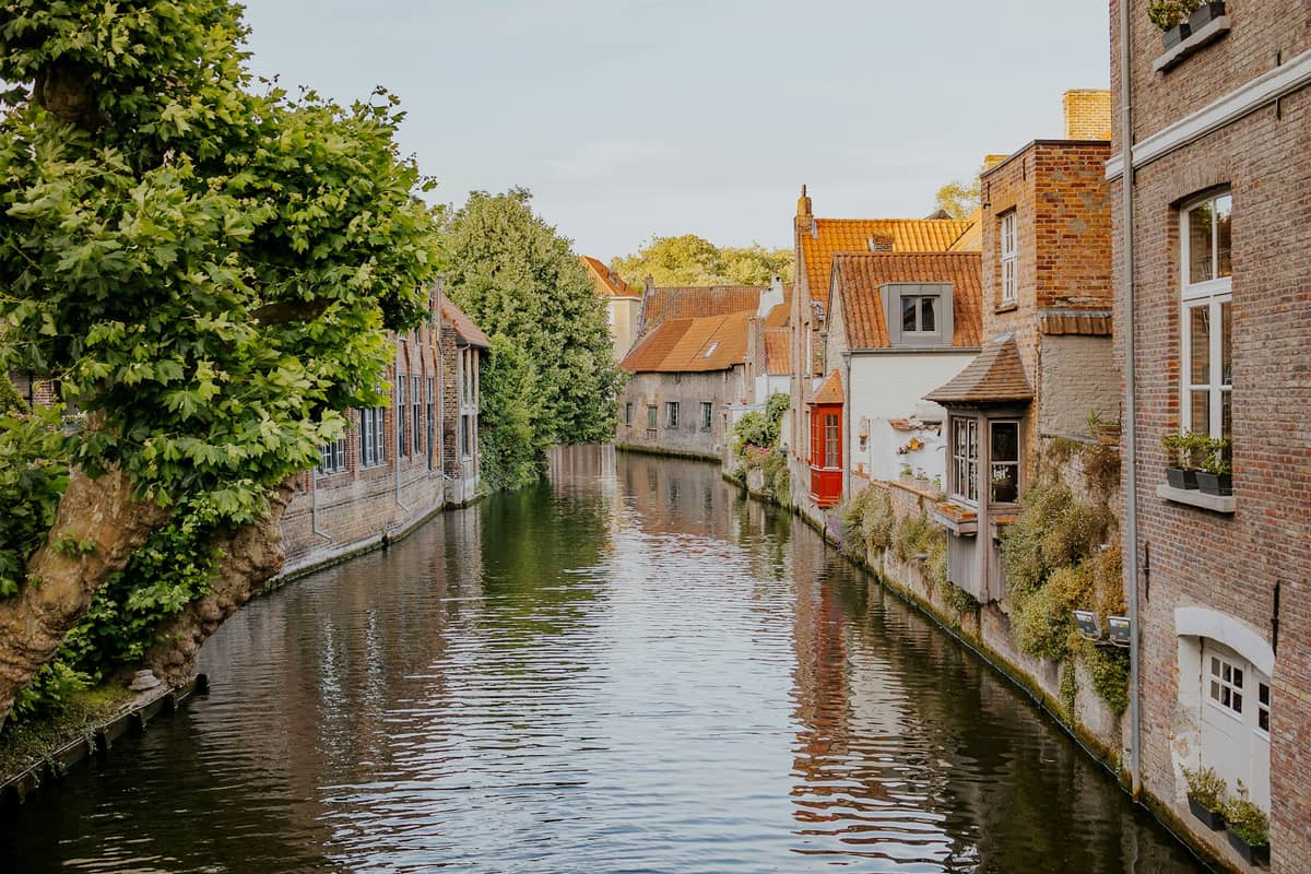 Quiet canal in Bruges lined with historic brick houses and lush trees, showing a peaceful, lesser-known side of the city away from tourist crowds.