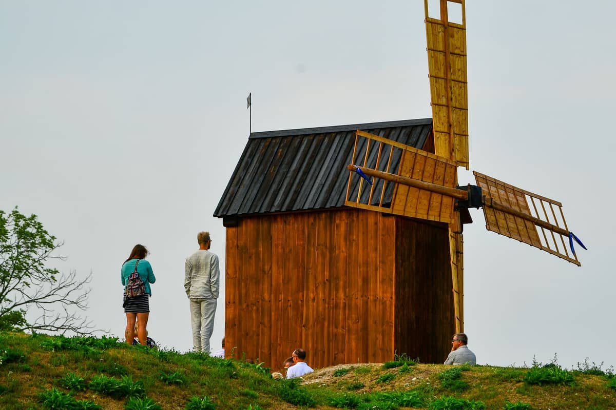 Small wooden windmill atop a grassy rampart with people standing nearby under a cloudy sky.