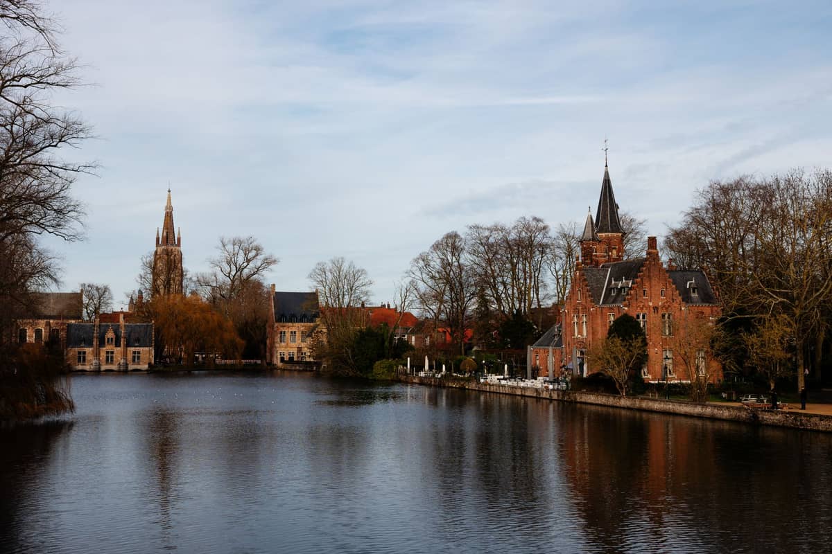 View of Bruges’ Minnewater Lake and historic brick buildings with bare winter trees and medieval spires reflecting in the calm water.