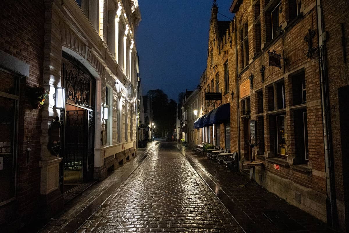 A quiet cobblestone street in Bruges' city center at night, glistening with wetness and lined with historic brick buildings.