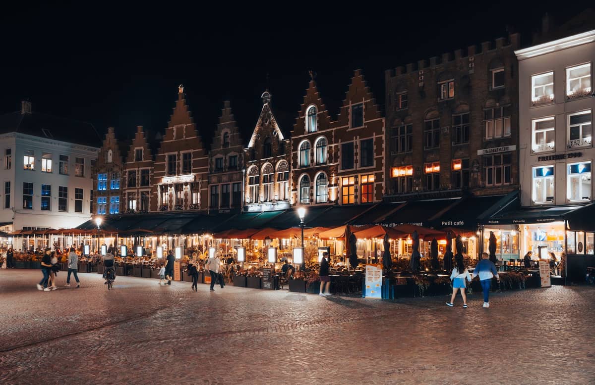 Night view of Markt Square in Bruges, with people dining at brightly lit outdoor restaurants beneath traditional stepped-gable facades.