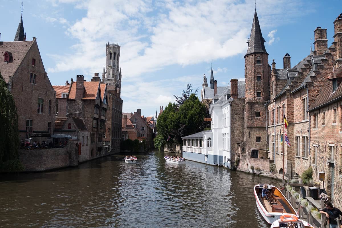 Classic view of Bruges' Rozenhoedkaai canal corner featuring medieval brick buildings, boats on the water, and the Belfry tower framed in the background.