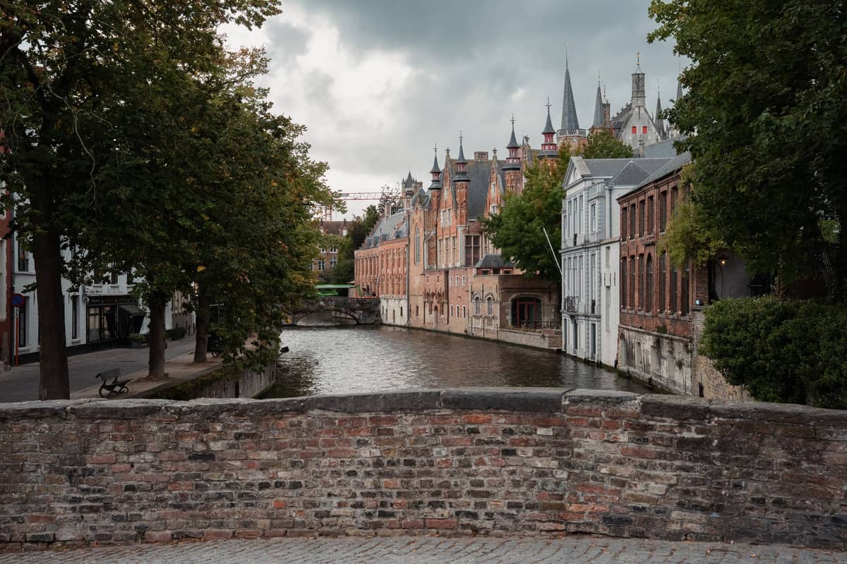 Classic medieval canal scene in Bruges with historic brick buildings, tree-lined waterway, and view toward city spires under a cloudy sky.
