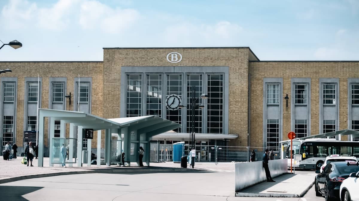 Exterior view of Bruges train station with brick facade, large clock, bus stop canopy, and people outside on a clear day