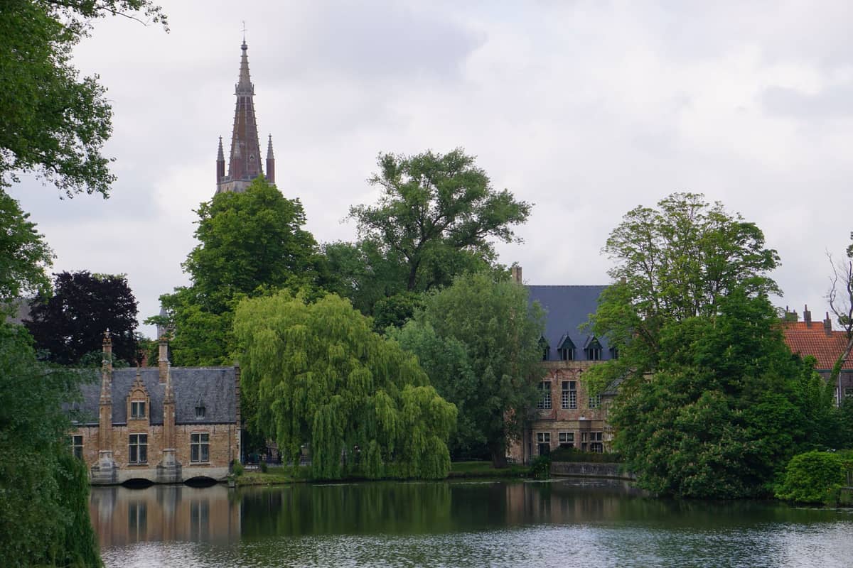 The Minnewater lake in Bruges with historical buildings, willow trees, and the spire of a church in the background.