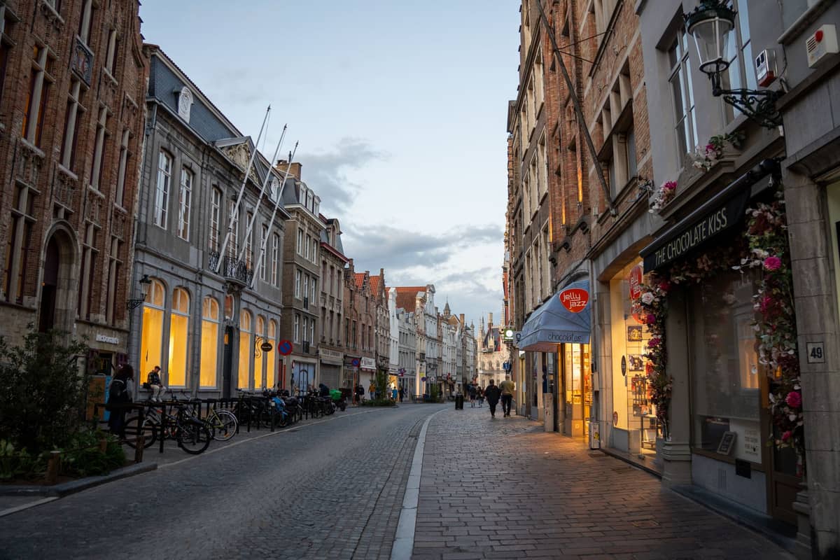 Pedestrian street in Bruges lined with boutique stores and warm lights, with people strolling along cobblestones at dusk.