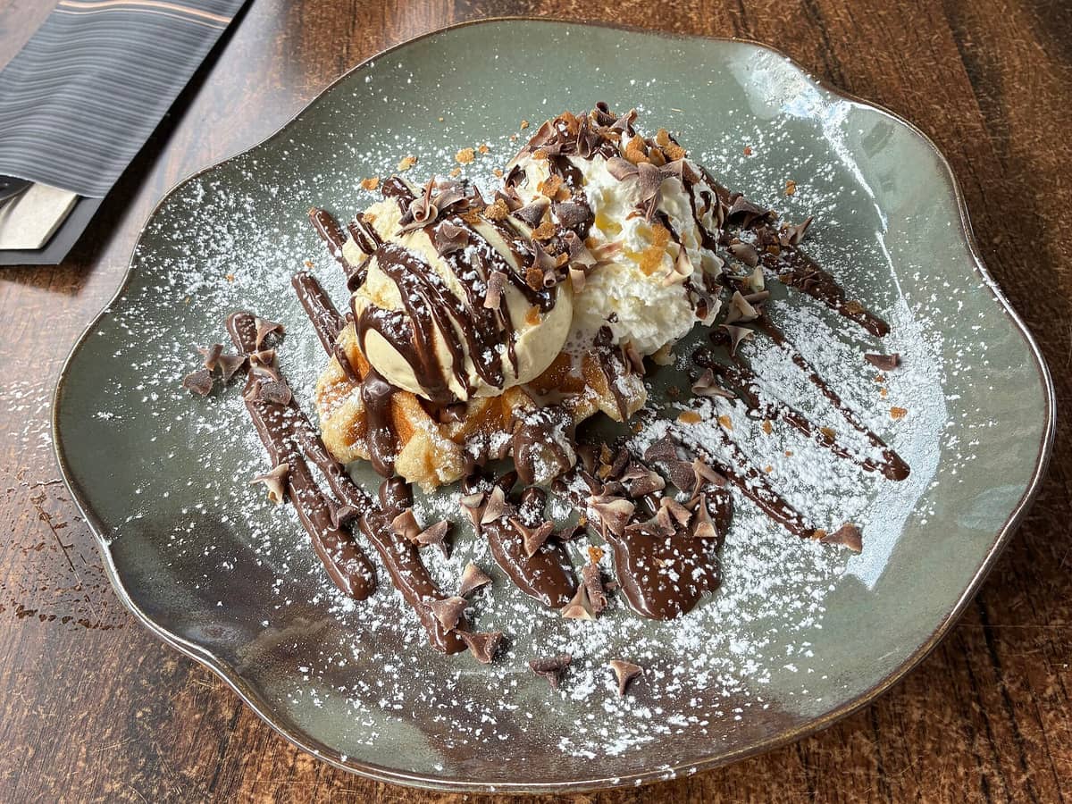 A close-up of a Belgian waffle in Bruges topped with whipped cream, chocolate sauce, vanilla ice cream, and chocolate shavings on a rustic plate.