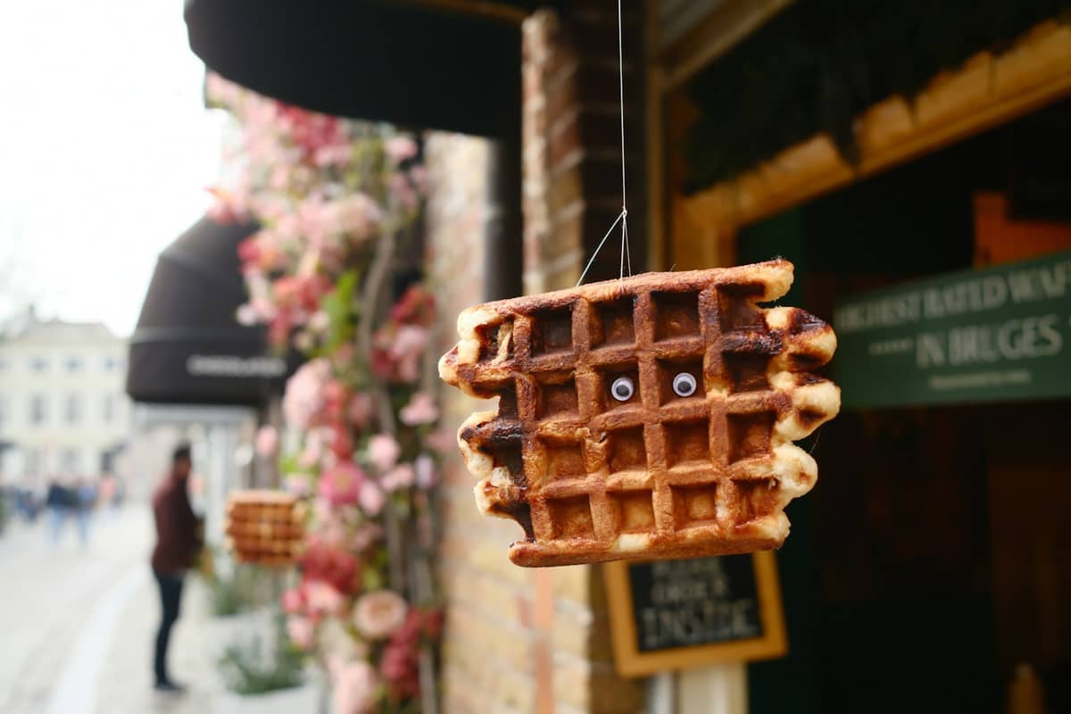 Close-up of a Belgian waffle with googly eyes hanging outside a Bruges waffle shop, floral decorations and a city street in the background.