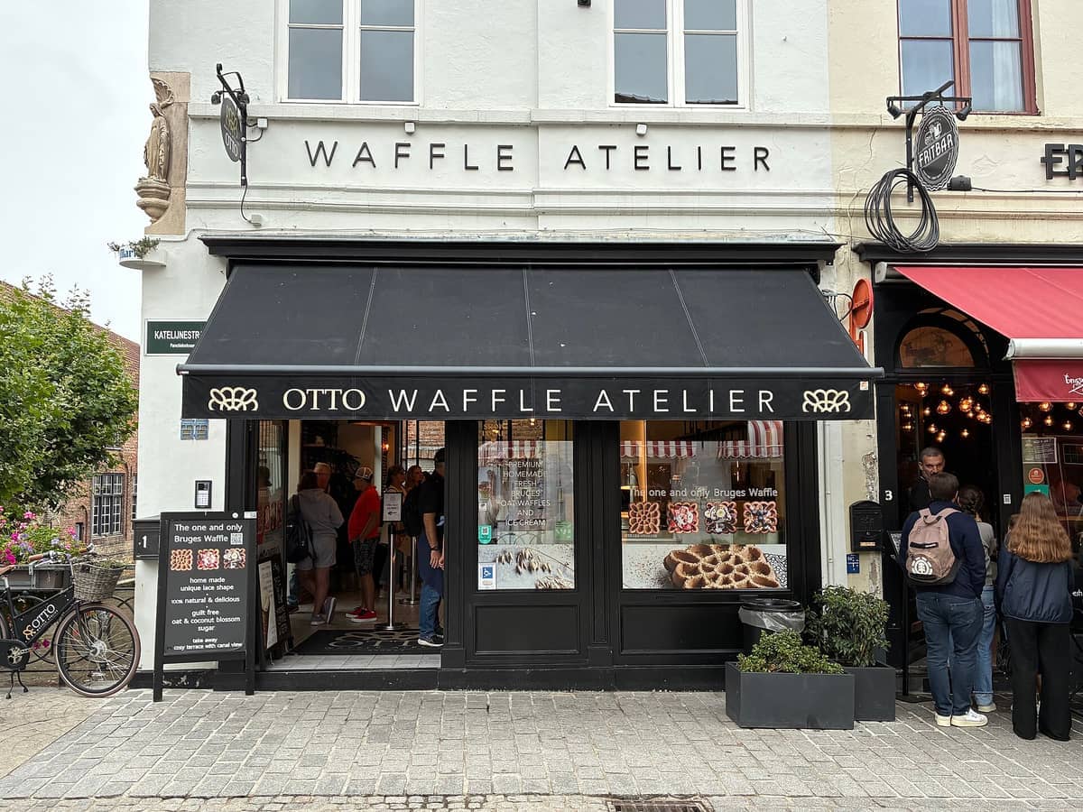 Street view of OTTO Waffle Atelier in central Bruges, with people waiting outside the black awning storefront on a cloudy day.