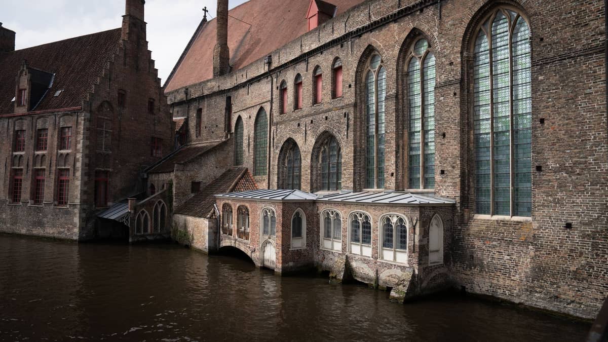 View of the exterior of the historic Groeningemuseum on Dijver canal in Bruges, with large arched windows and brick facade directly beside the water.