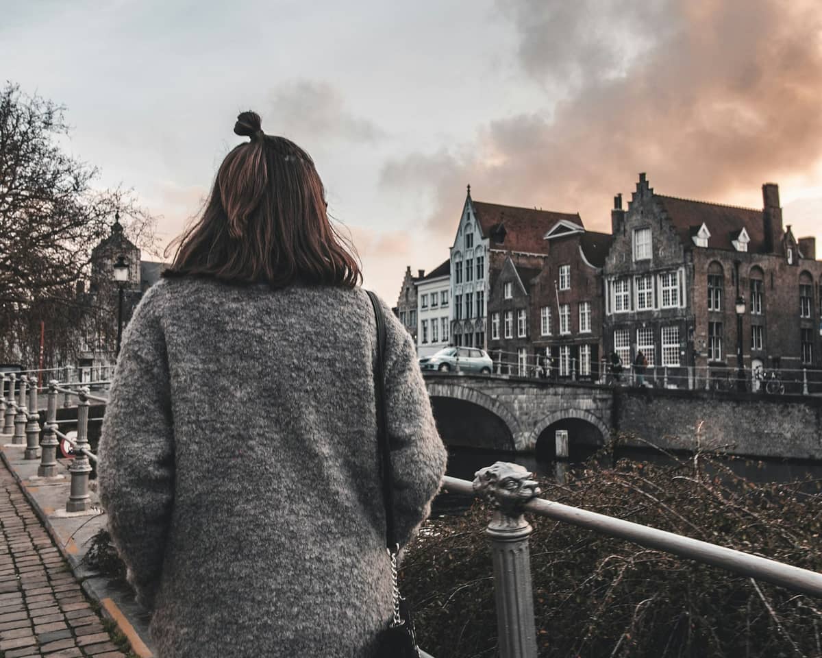 Woman in a gray coat walking along a cobblestone canal path in Bruges, with medieval Flemish buildings and an arched bridge at sunset.