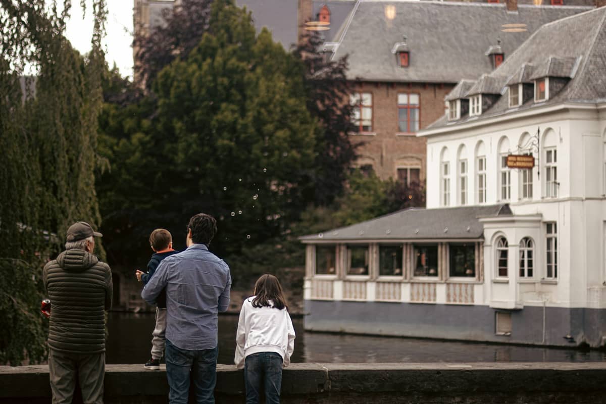 A family stands by a canal in Bruges, Belgium, overlooking historic buildings including a white riverside restaurant with large windows.