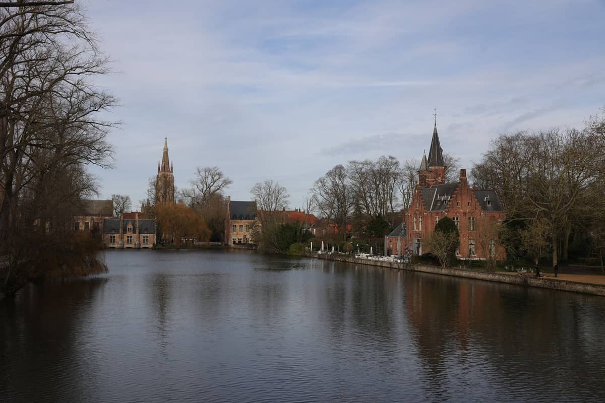 View of Minnewaterpark in Bruges with a large pond, historic buildings, and tree-lined banks, under a blue sky.