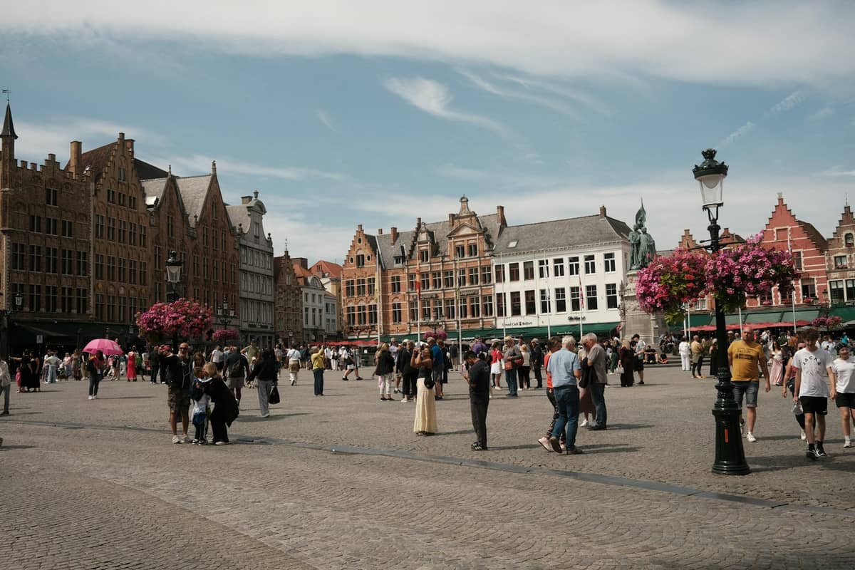 Wide view of Bruges main square with families and tourists walking among historic stepped-roof buildings on a sunny day in late spring or summer.