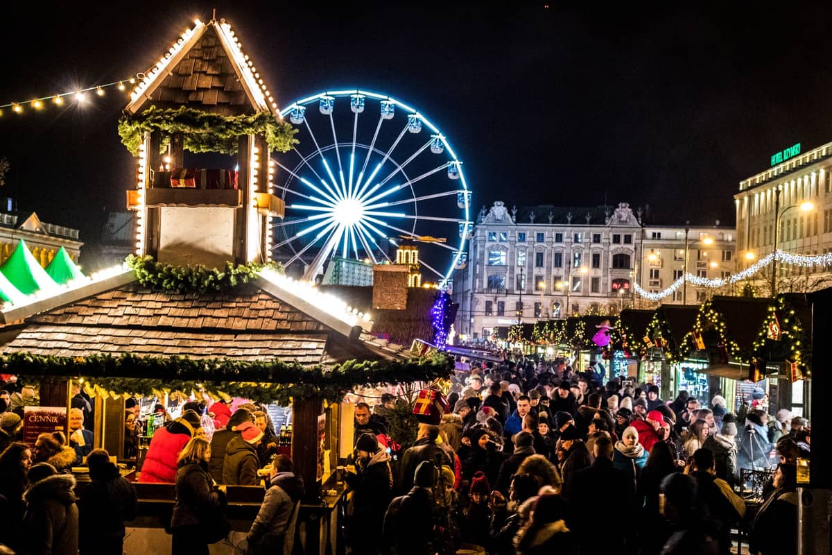 Grandes multidões no mercado de Natal de Bruxelas com luzes festivas, bancas de mercado e uma roda-gigante à noite.
