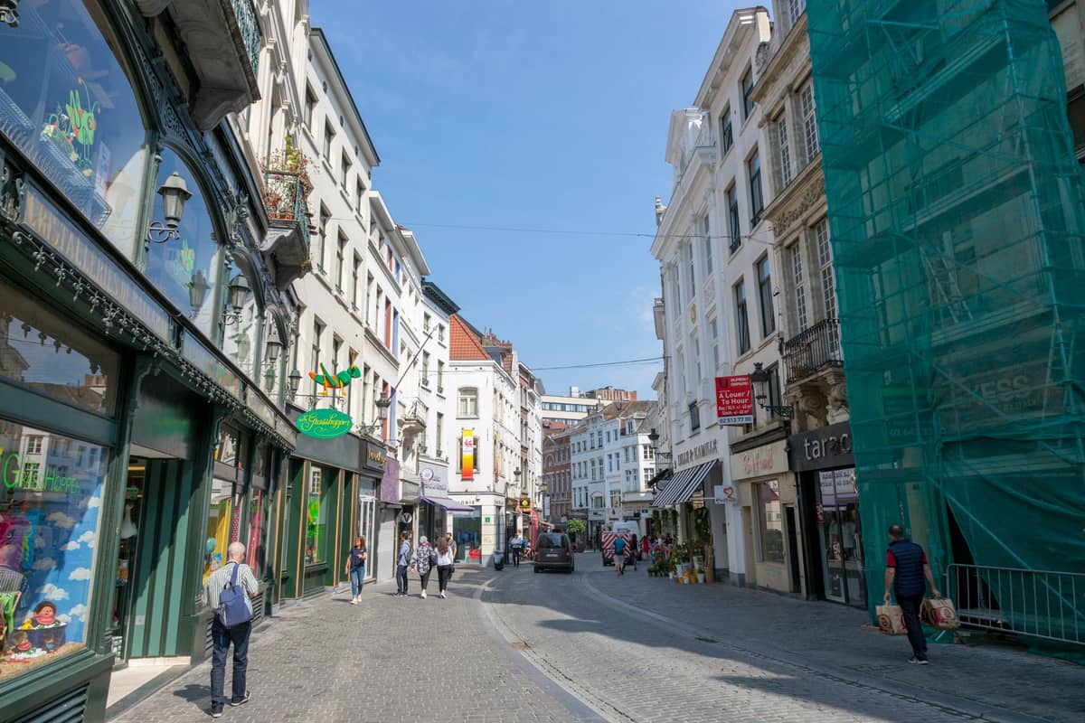 Uma animada cena de rua no centro de Bruxelas com lojas, pedestres e arquitetura clássica da cidade sob um céu azul.