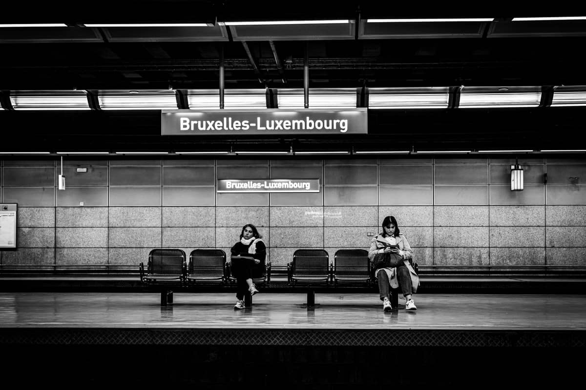 Duas pessoas esperam em bancos na plataforma da estação de metrô Bruxelles-Luxembourg, com a placa da estação acima delas.