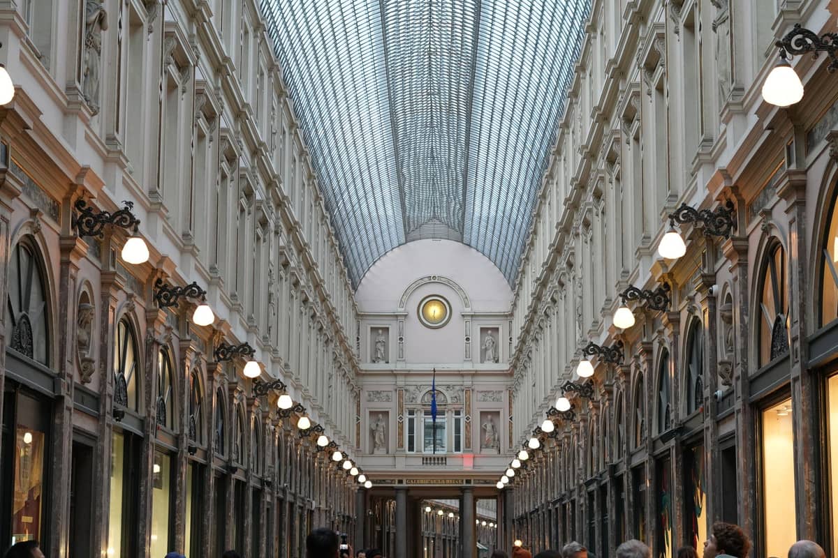 Vista ampla das Galeries Royales Saint-Hubert em Bruxelas, com teto de vidro, arquitetura ornamentada, lojas e pessoas caminhando no interior.