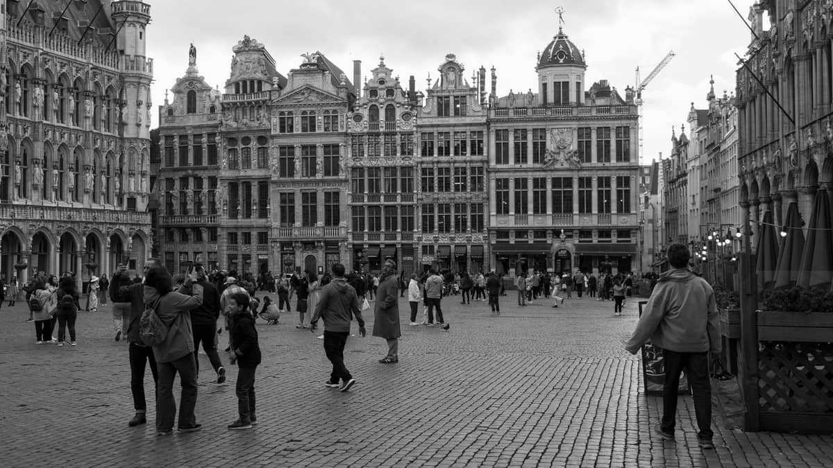 Foto em preto e branco de pessoas passeando e tirando fotos na Grand Place de Bruxelas, rodeada por ornamentados edifícios históricos.