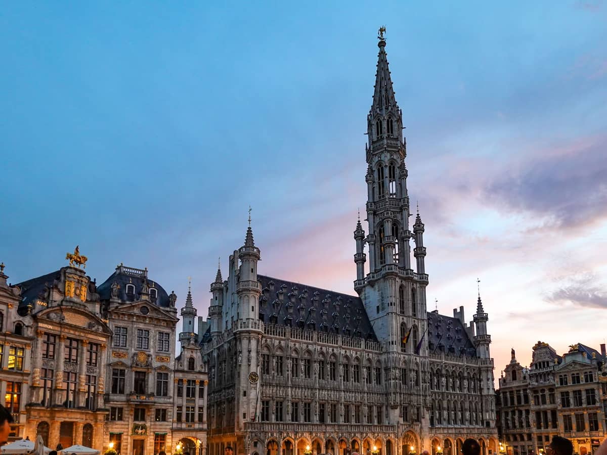 Vista ao entardecer da Grand Place em Bruxelas com ornamentados edifícios históricos e a torre iluminada da Câmara Municipal sob um céu rosa e azul.
