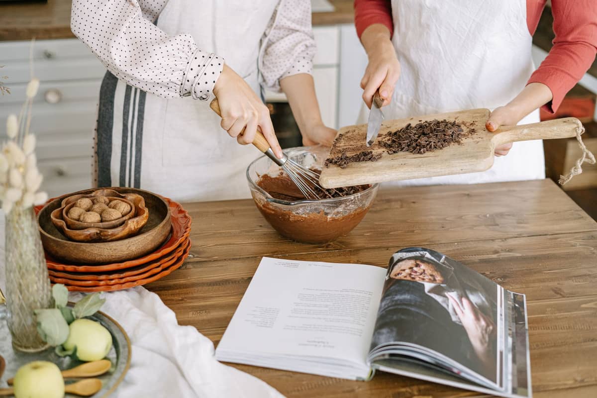 Duas pessoas com aventais preparando chocolate juntas em uma mesa de cozinha com um livro de receitas aberto e tigelas de ingredientes.