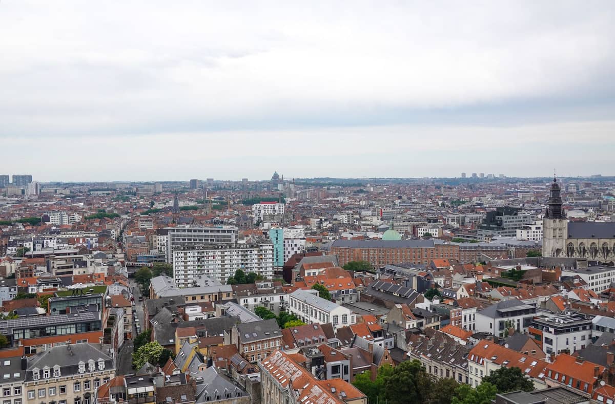 Vista urbana ampla sobre Bruxelas com telhados, edifícios modernos e históricos espalhados e a cidade se estendendo ao longe sob um céu nublado.