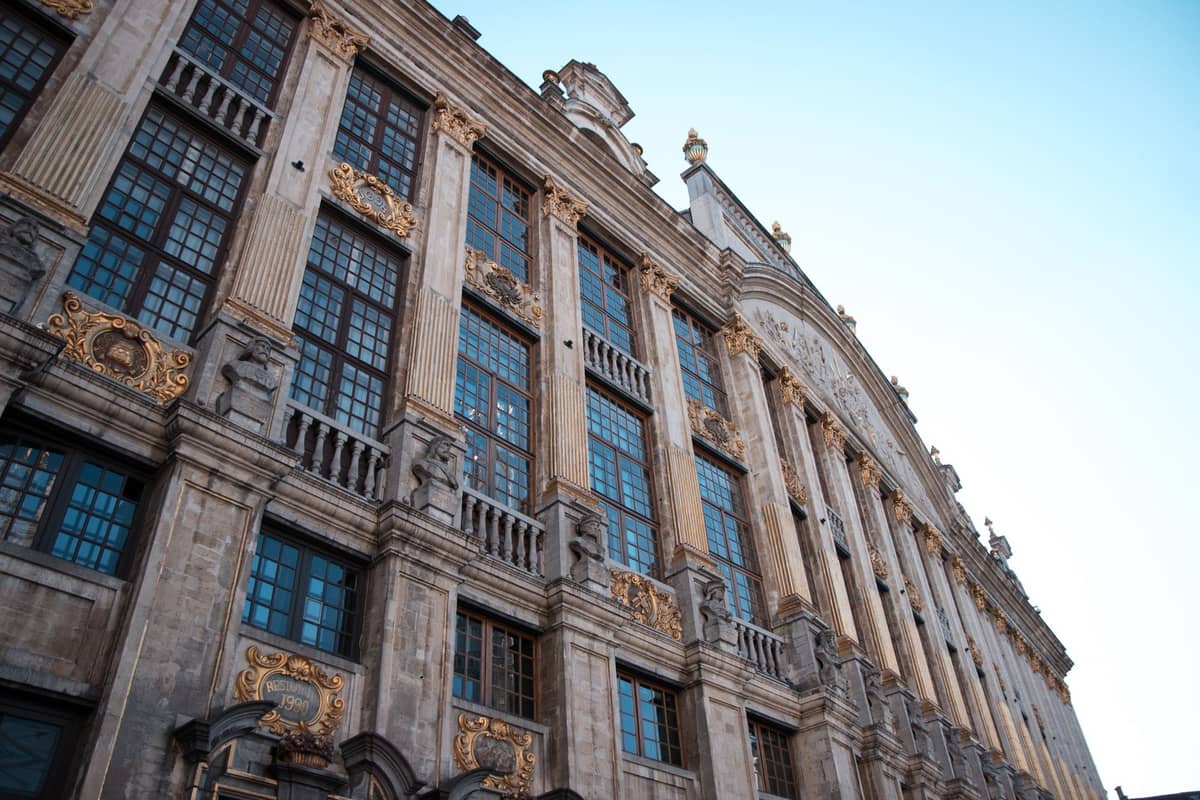 Grande edifício histórico com fachada ornamentada e detalhes dourados em Bruxelas, fotografado ao nível da rua contra um céu azul limpo.