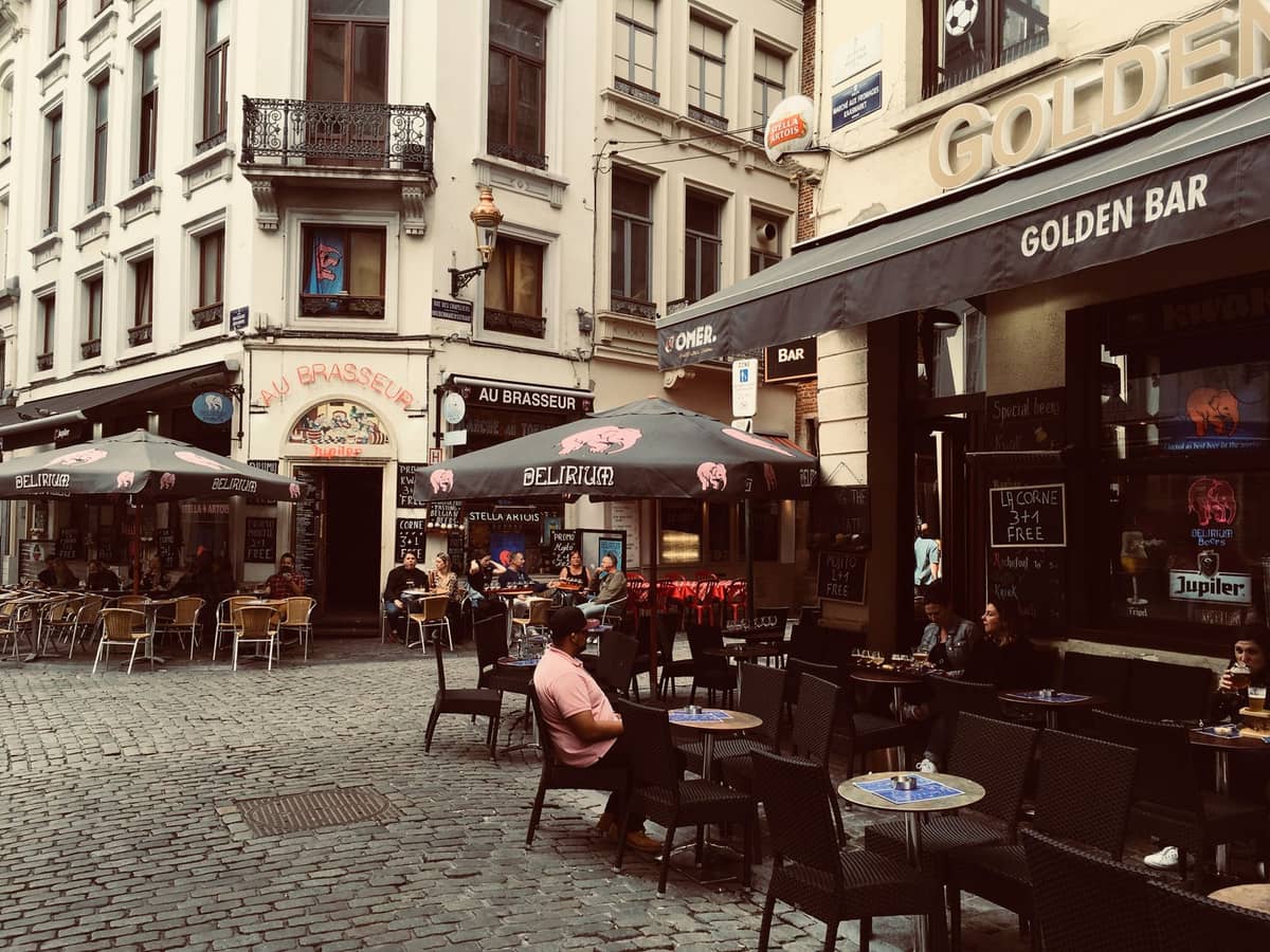 Terraços de cafés e bares em uma rua de paralelepípedos no centro de Bruxelas, com pessoas aproveitando drinks e a vida noturna local.