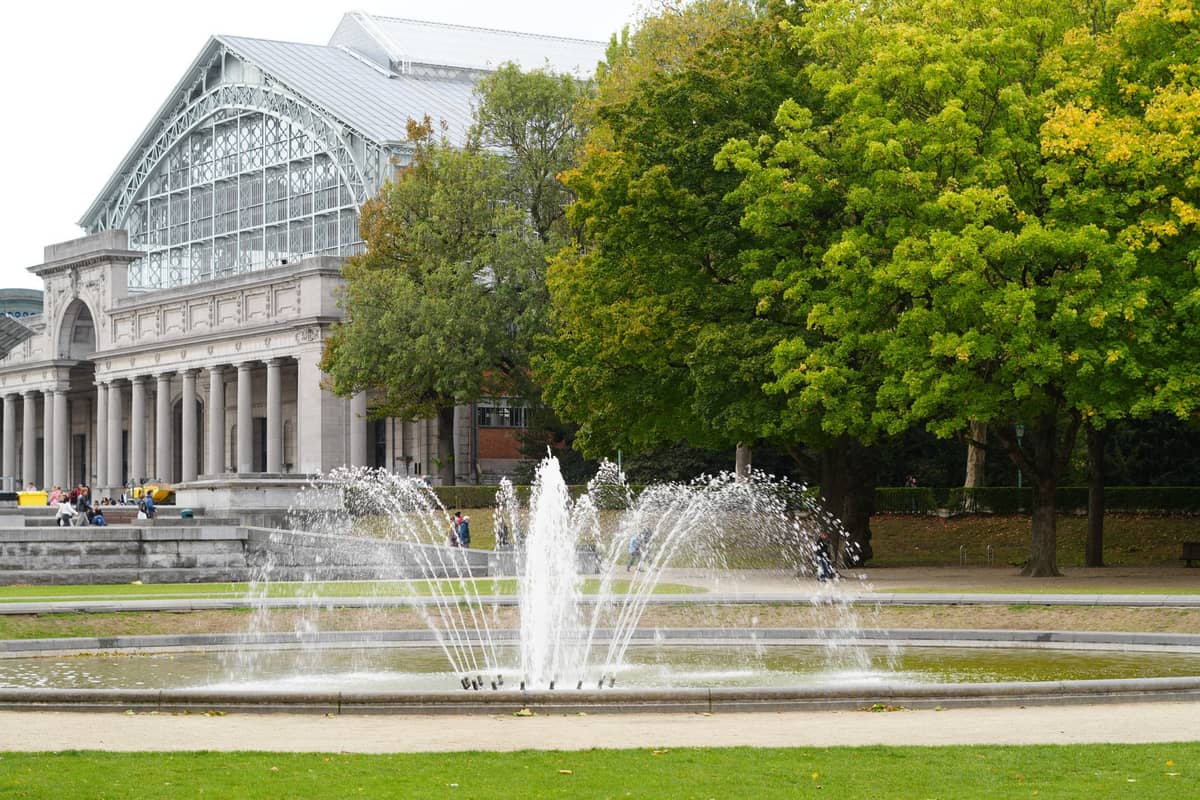 Vista panorâmica do Parc du Cinquantenaire e do Museu de Arte e História em Bruxelas, com uma fonte e árvores verdes em primeiro plano.