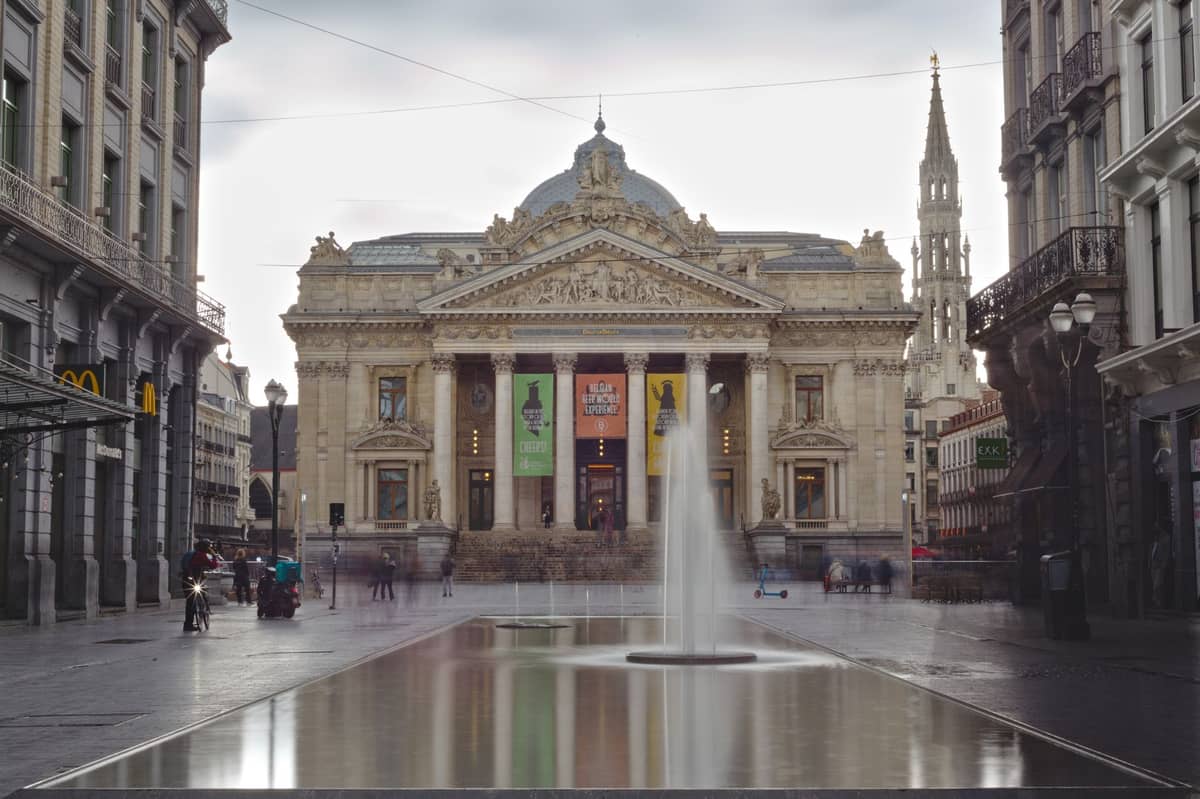 Vista frontal da Bolsa de Valores de Bruxelas com uma fonte em primeiro plano, pessoas caminhando e céu nublado ao fundo.