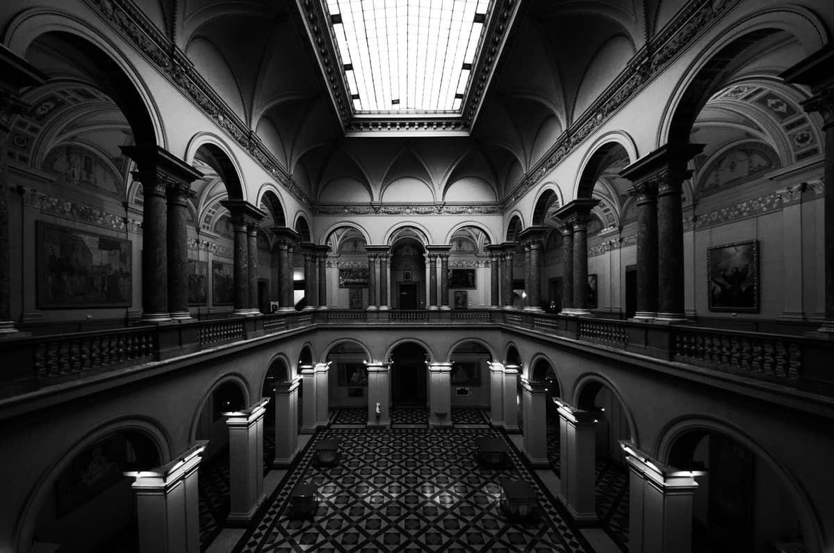 Grande salão interior clássico com arcos, colunas, galeria superior, claraboia e pinturas nas paredes em um espaço de tipo grande museu.