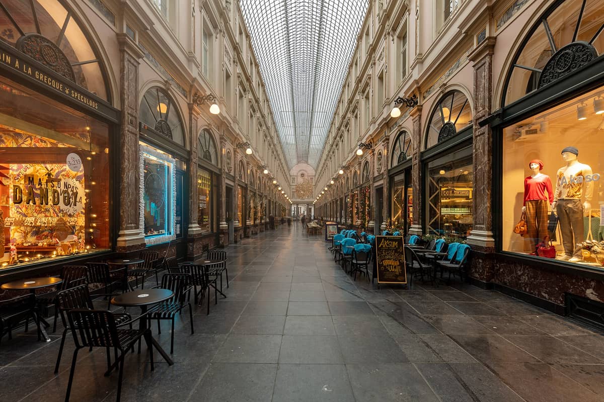 Vista ampla das Galeries Royales Saint-Hubert em Bruxelas, mostrando elegante arquitetura histórica, telhado de vidro, lojas e cafés na calçada sob cobertura.