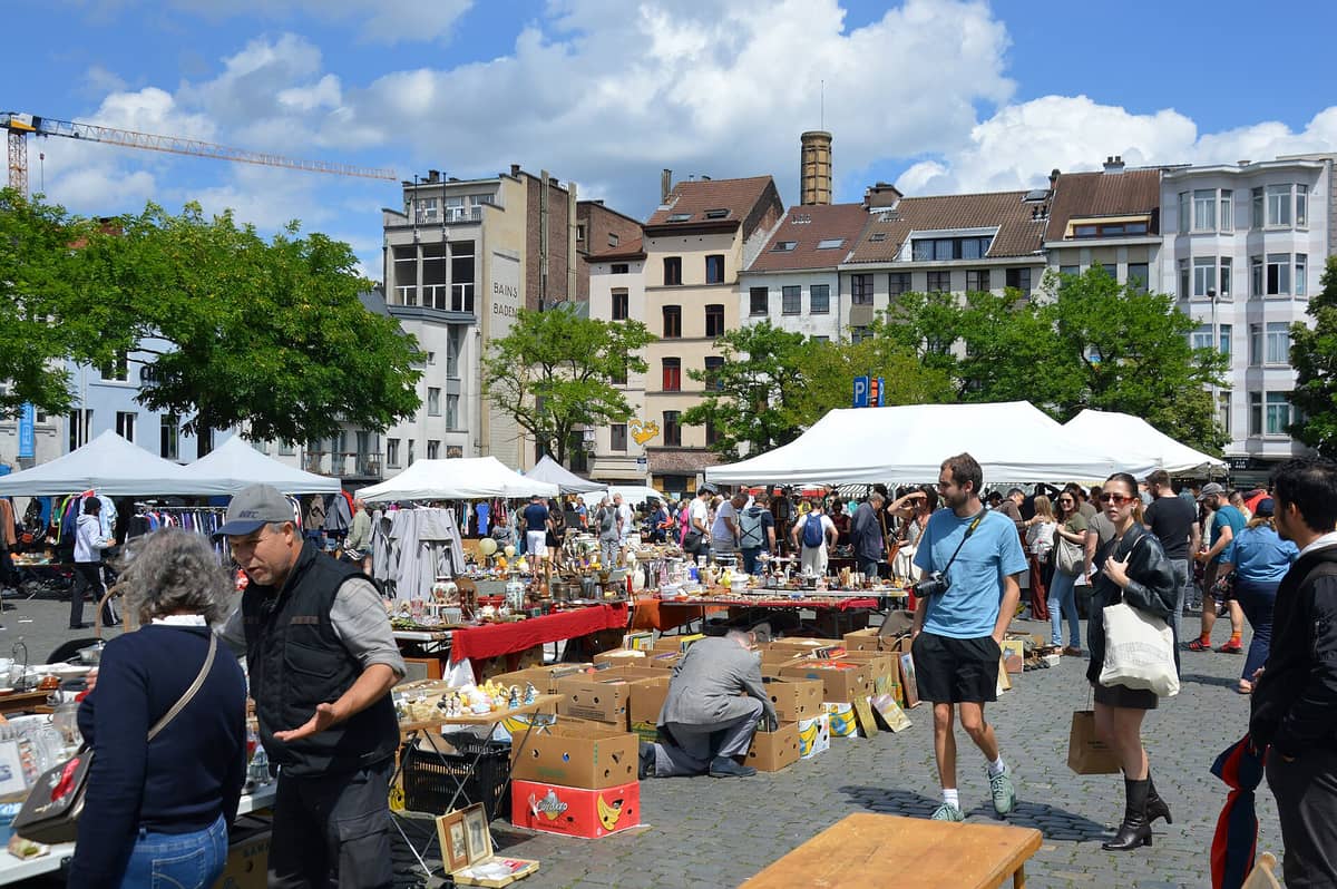 Movimentado mercado de pulgas no bairro de Marolles em Bruxelas com pessoas a explorar mesas de antiguidades e bugigangas, situado numa praça ao ar livre com edifícios da cidade ao fundo.