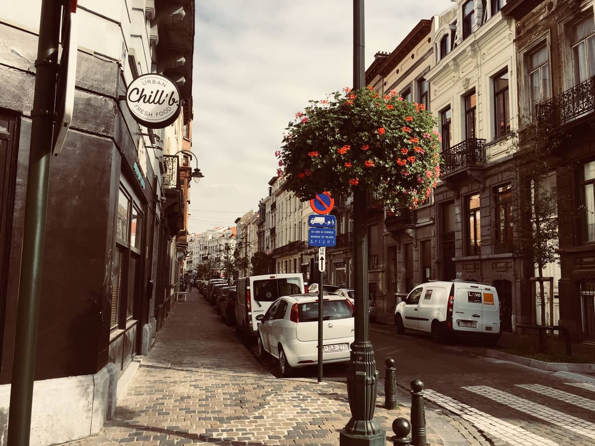 Encantadora rua de Bruxelas com boutiques, carros estacionados, fachadas históricas e um cesto de flores pendurado, evocando o ambiente de compras boutique de Dansaert.
