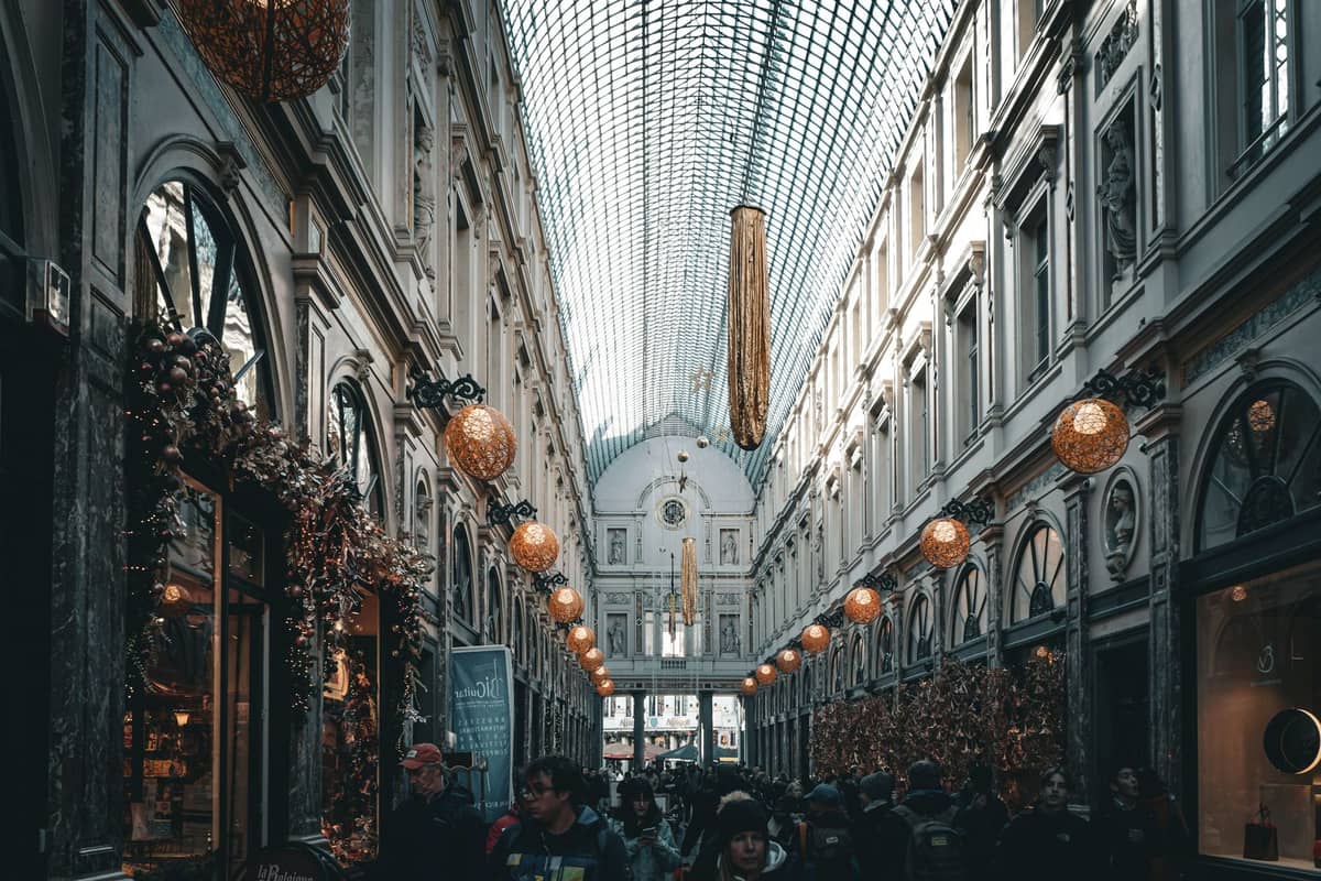 Vista interna das Galeries Royales Saint-Hubert em Bruxelas, com pessoas fazendo compras, luzes decorativas suspensas e teto abobadado de vidro.