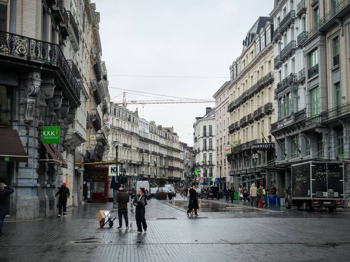 Rua pedonal em Bruxelas ladeada por lojas de redes, grandes letreiros comerciais e pessoas a fazer compras num dia nublado.