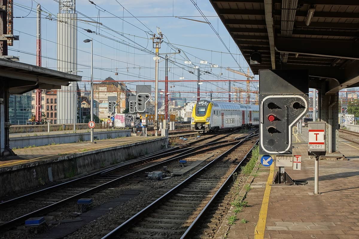 Um trem Eurocity de dois andares da Belgian Rail chegando nas trilhas externas da estação Bruxelles-Nord com edifícios da cidade ao fundo.