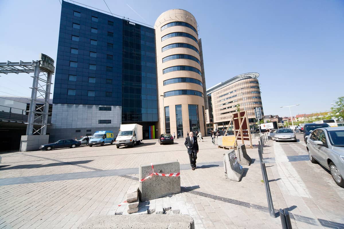 Vista ao nível da rua do moderno exterior da estação ferroviária de Bruxelas-Midi com edifícios de escritórios ao redor e pessoas caminhando em um dia ensolarado.