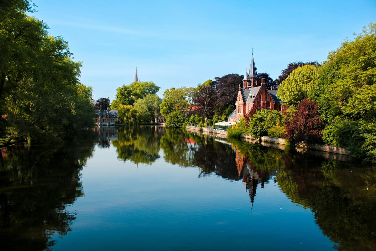Vista do Lago Minnewater e do charmoso castelo de tijolos em Bruges, rodeado por árvores exuberantes e refletido nas águas tranquilas.