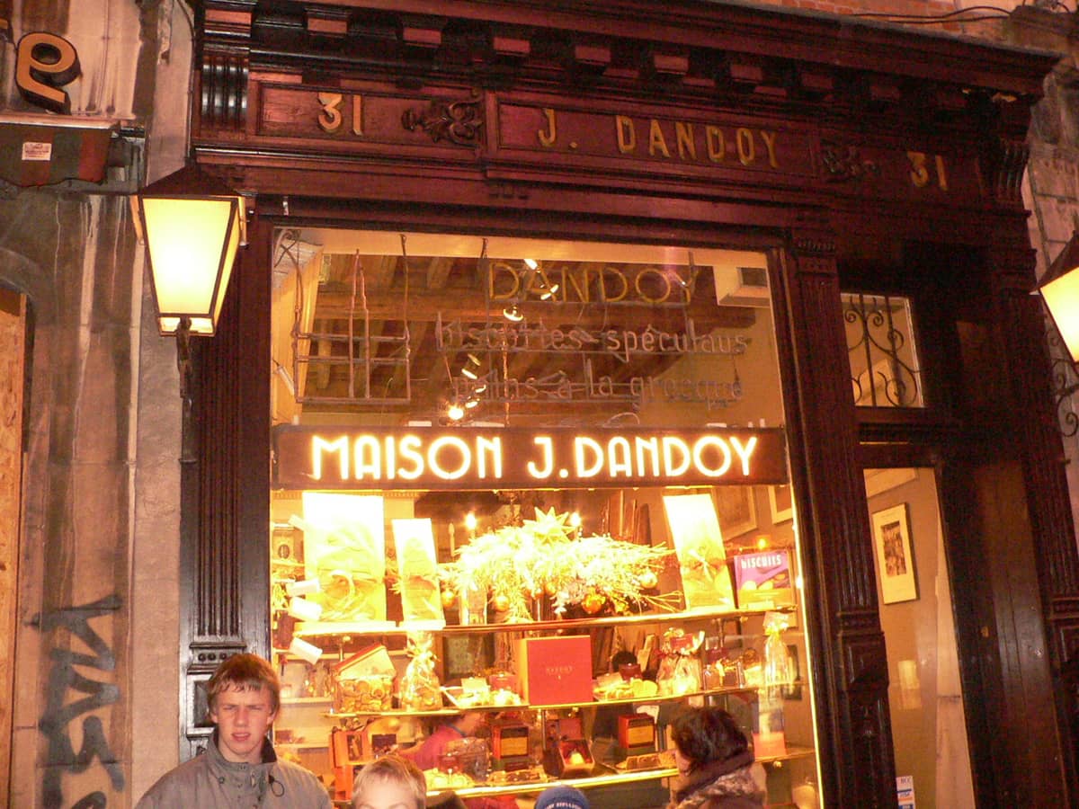 Exterior da padaria Maison J. Dandoy em Bruxelas à noite, bem iluminada com sua placa visível acima da vitrine e pessoas em frente.