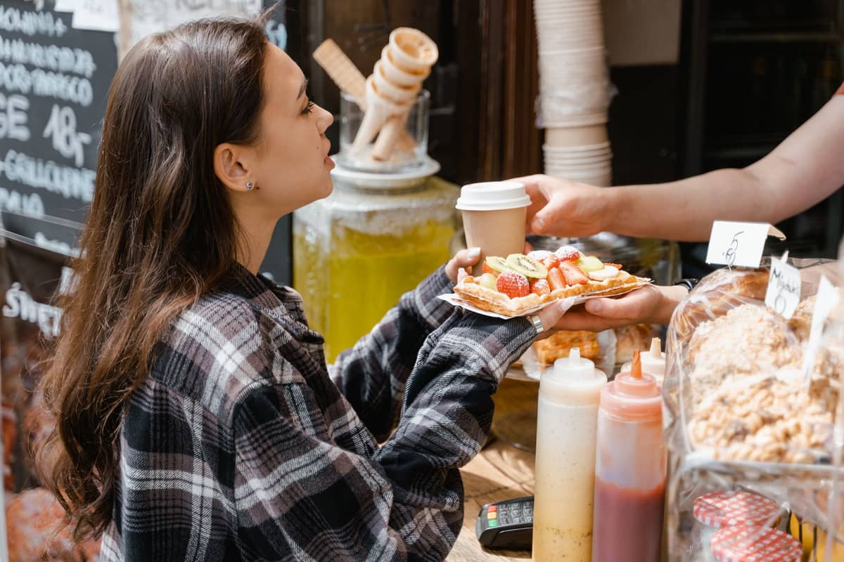 Mulher recebendo um waffle coberto com frutas e um café em uma barraca de rua, com vários ingredientes em primeiro plano