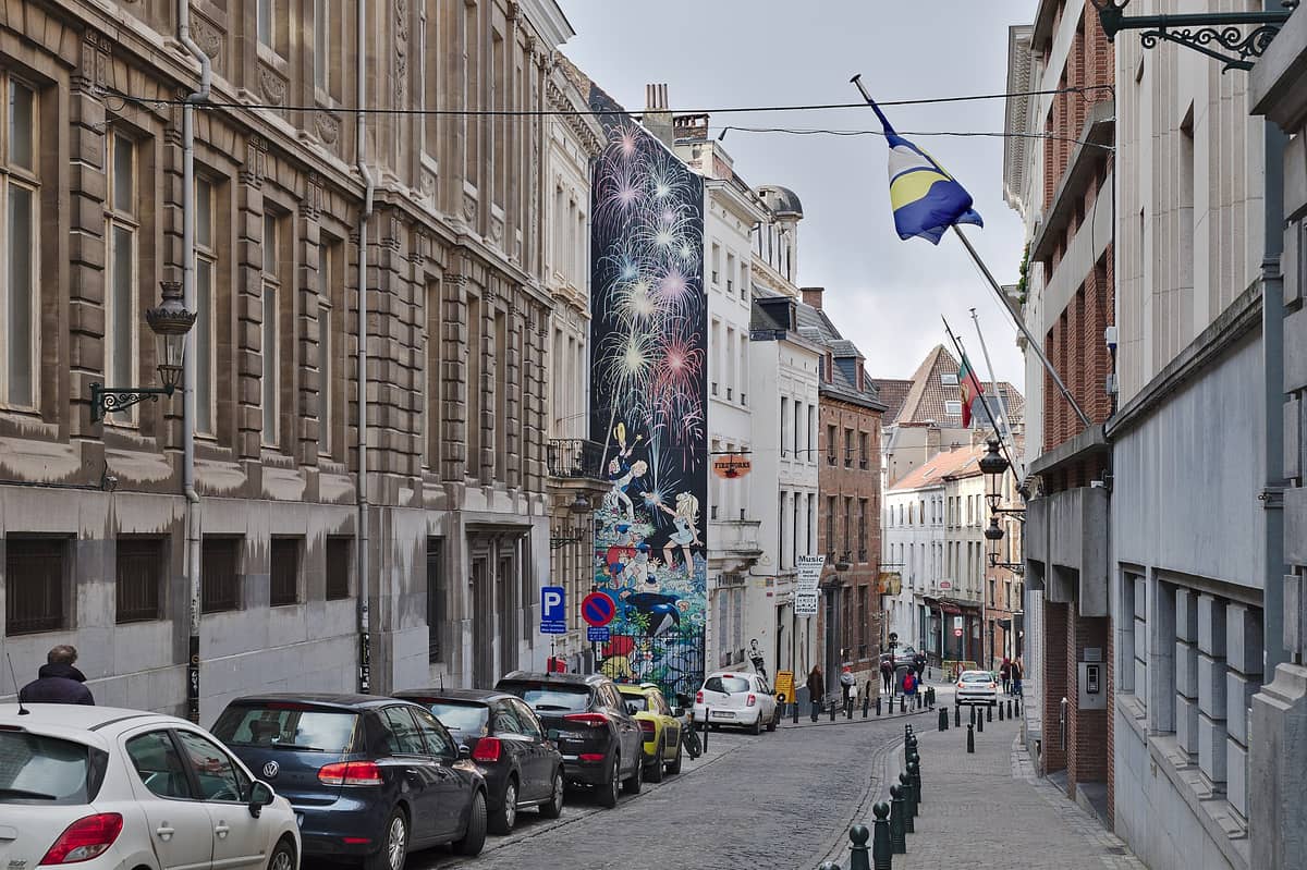 Uma rua de paralelepípedos em Bruxelas com carros estacionados e um grande mural de banda desenhada com personagens coloridos e fogos de artifício pintados na lateral de um edifício.