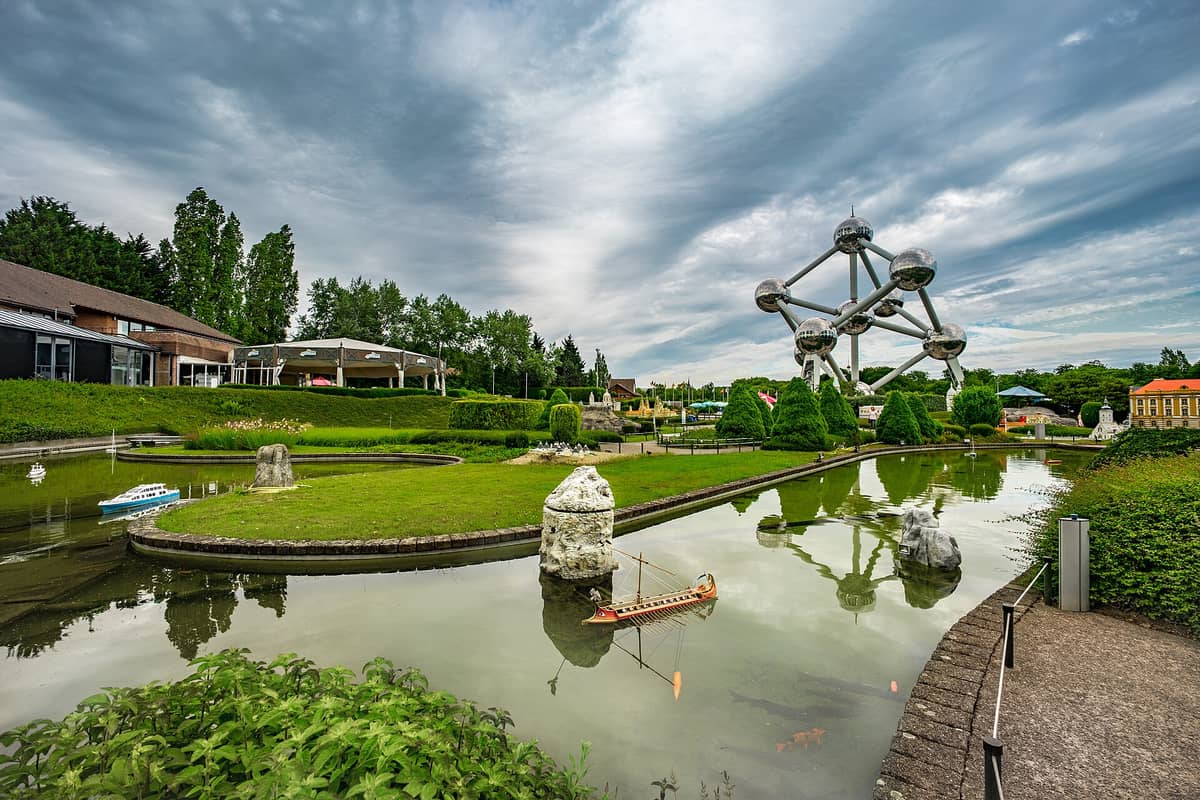 Vista ampla do parque Mini-Europa de Bruxelas com pequenos barcos em miniatura, vegetação exuberante e o Atomium ao fundo sob um céu nublado dramático.