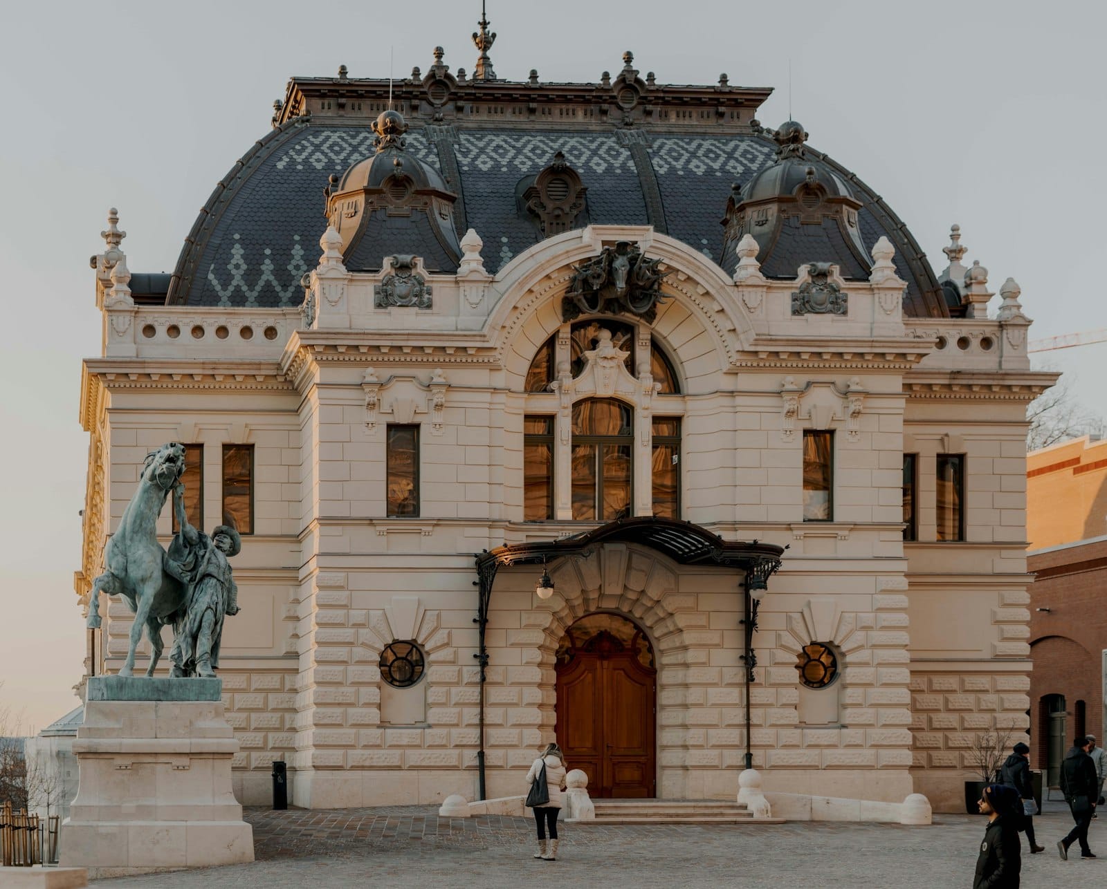 Budapest'te fotoğraflanmış, kubbeli çatısı, işlemeli pencereleri ve önünde at heykeli bulunan zarif ve süslü Art Nouveau binası.