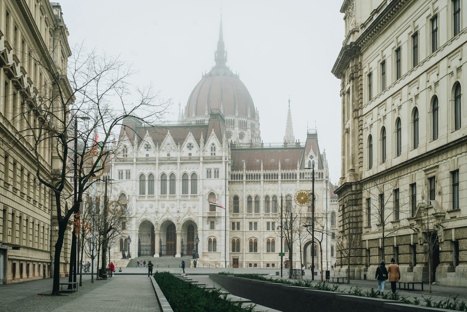 Breite Straße mit Fußgängern, die auf das imposante ungarische Parlamentsgebäude in Budapest zuführt, flankiert von klassischer Architektur auf beiden Seiten im sanften Tageslicht.
