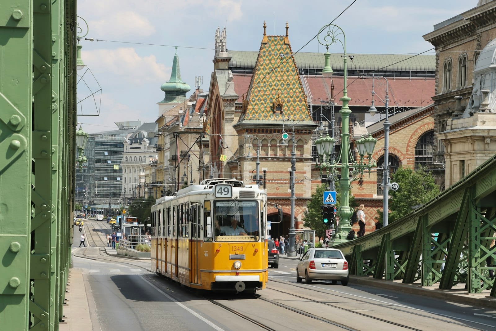 Gelbe Budapester Straßenbahn überquert die Freiheitsbrücke mit der Großen Markthalle und Stadtsehenswürdigkeiten im Hintergrund an einem sonnigen Tag.